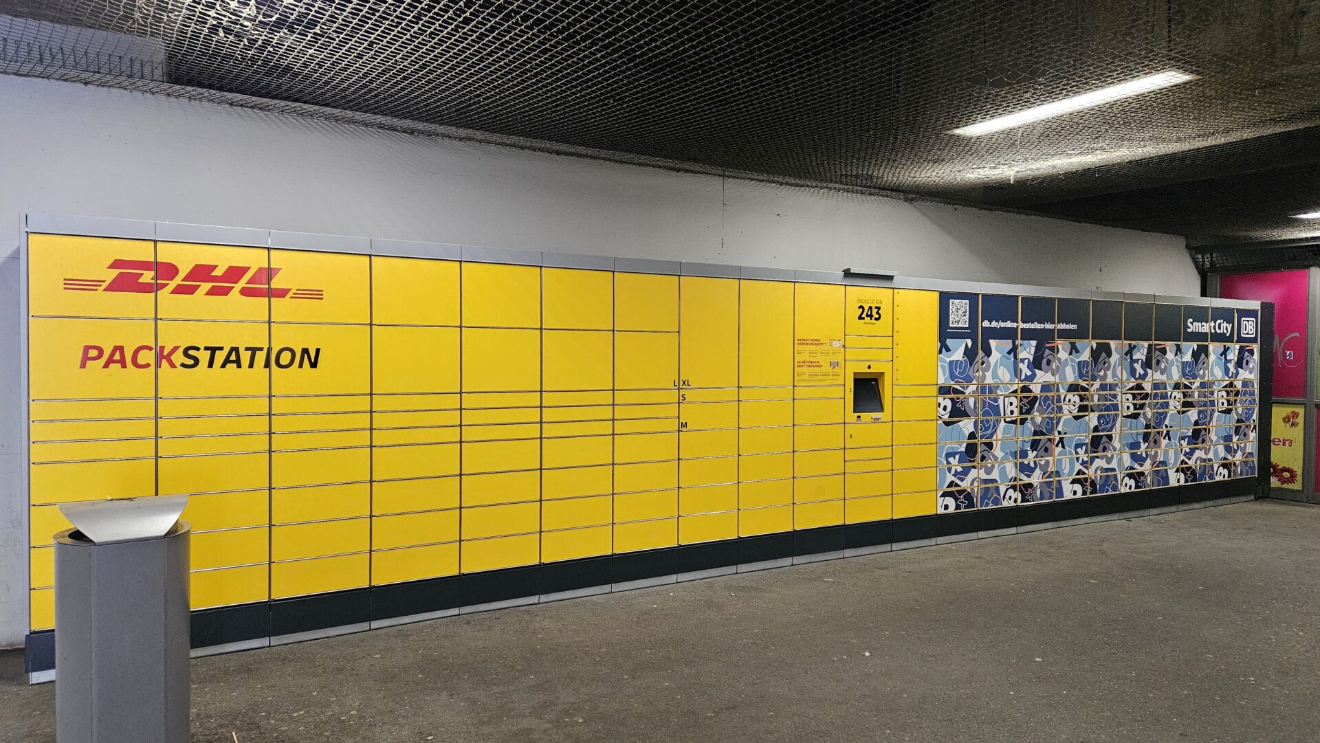 Gelbe DHL Packstation am Bahnhof Mitte Dresden, aufgestellt neben Fahrradständern und unter einer Überdachung, bereit zur Paketabholung.