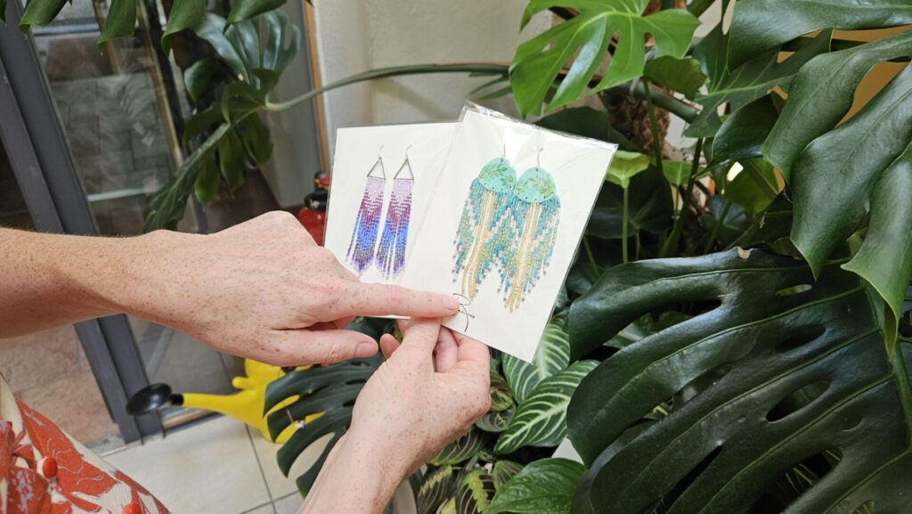 Racine Morgan in einem Sommerkleid, lächelnd, vor tropischen Pflanzen im Café Almuth. Sie hält drei Paar selbst gestaltete Ohrringe aus Modelliermasse in der Hand.